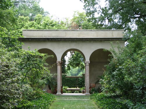 A serene garden scene featuring a stone archway with columns, surrounded by lush greenery and trees.