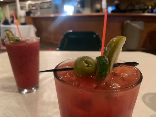 Two cocktails with garnishes of olives, lime, and a straw, set on a table in a dimly lit bar.