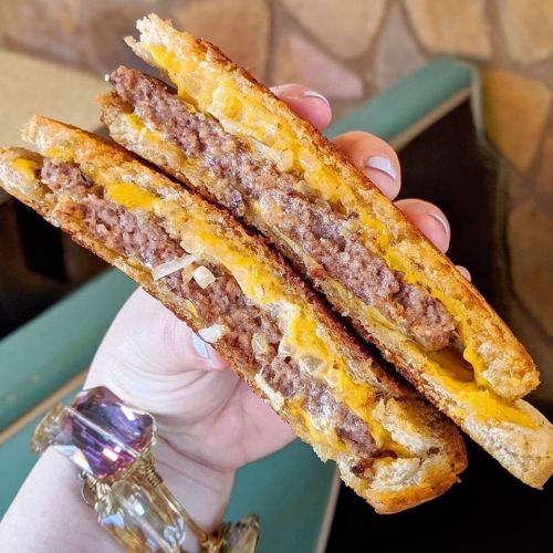 A hand holding a grilled cheese sandwich filled with beef patty and melted cheddar cheese, showcasing its layers.