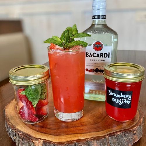 A refreshing strawberry mojito in a glass, flanked by jars of fresh strawberries and a strawberry mojito mix on a wooden surface.