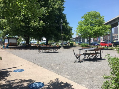 A sunny outdoor space with tables and chairs surrounded by trees and a gravel path, under a clear blue sky.