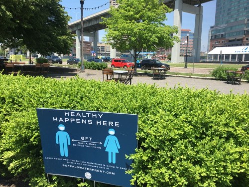 A sign promoting health guidelines stands in a park with tables, greenery, and a view of a nearby road and buildings.