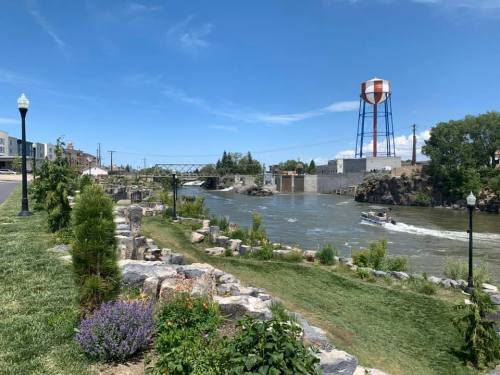 A scenic river view with greenery, a water tower, and a boat passing by under a clear blue sky.