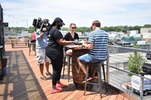 A woman serves food to a man at a rooftop bar while a cameraman films the interaction.