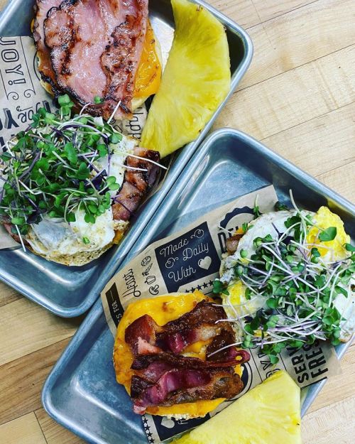Two trays of gourmet breakfast sandwiches topped with microgreens, bacon, and eggs, served with pineapple slices.
