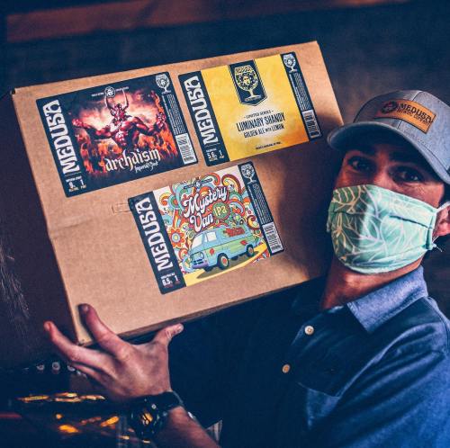 A person wearing a mask holds a box featuring colorful beer labels, including "Mystery Day" and "Luminous Savant."