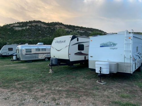 A row of parked campers and trailers on grassy land with a mountainous backdrop under a cloudy sky.