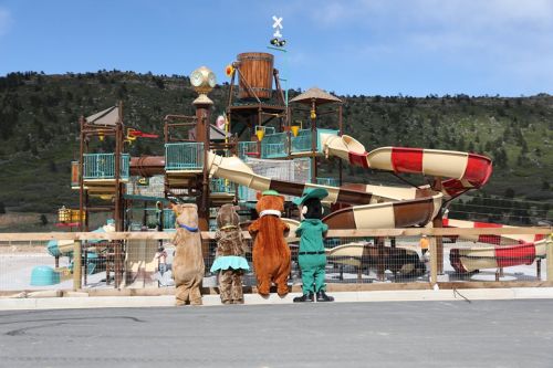 Three costumed characters stand in front of a colorful water slide playground against a scenic mountain backdrop.