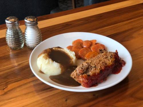 A plate of meatloaf with gravy, mashed potatoes, and glazed carrots, set on a wooden table with salt and pepper shakers.