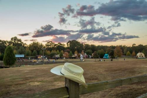 A white cowboy hat rests on a wooden fence, overlooking a scenic field at sunset with colorful clouds.