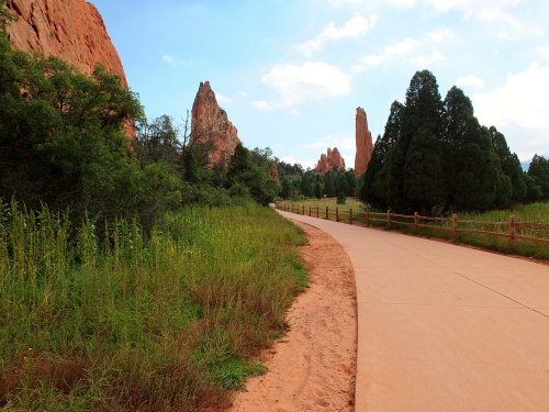A winding path through lush greenery, flanked by towering red rock formations and pine trees under a blue sky.