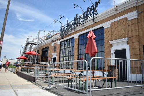 Exterior of a restaurant with large windows, outdoor seating, red umbrellas, and a decorative sign on a sunny day.