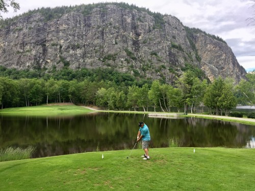 A golfer prepares to swing on a lush green course beside a calm lake, with a rocky mountain backdrop.