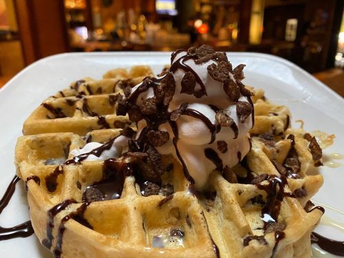 A close-up of a waffle topped with whipped cream, chocolate drizzle, and crunchy chocolate pieces on a white plate.