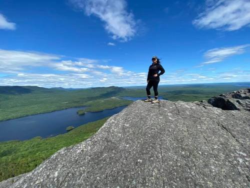 A person stands on a rocky mountain peak, overlooking a lush green landscape and a lake under a blue sky.