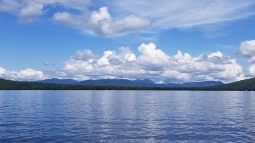 A serene lake reflects fluffy clouds and distant mountains under a bright blue sky.