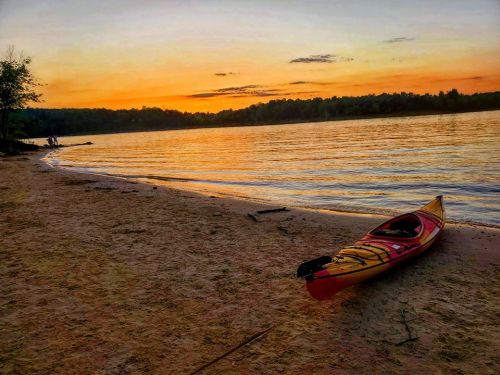 A colorful kayak rests on a sandy beach at sunset, with calm water and trees in the background.