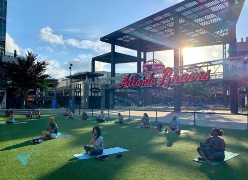 A group of people practicing yoga on a grassy area with a bright sun and the Atlanta Braves sign in the background.