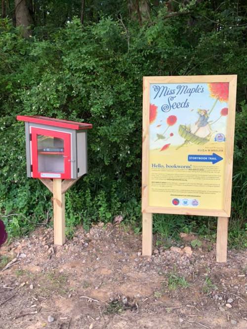 A small red and gray book box next to a sign for "Miss Maple's Seeds" on a wooded trail.
