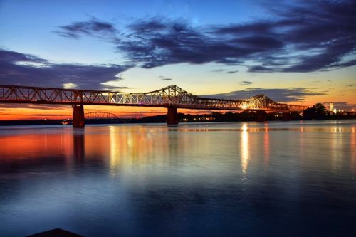 A serene sunset over a river, reflecting vibrant colors and a bridge silhouetted against the sky.