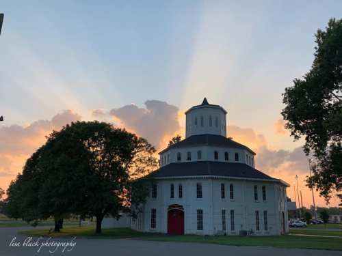 A round, white building with a dome, surrounded by trees, against a colorful sunset sky with rays of light.