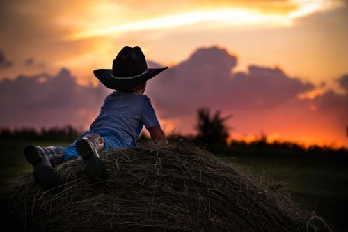 A child in a cowboy hat sits on a haystack, watching a colorful sunset with clouds in the background.