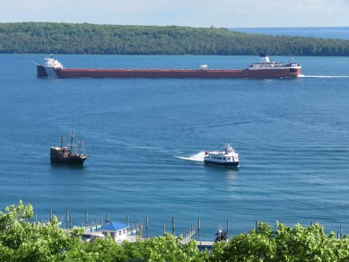 A large cargo ship and a smaller boat navigate a calm blue lake, with green trees in the background.
