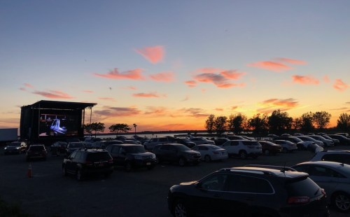 A sunset over a parking lot with cars, featuring a large outdoor screen displaying a movie.