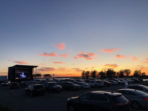 A sunset over a parking lot with cars, featuring a large screen displaying a performance.