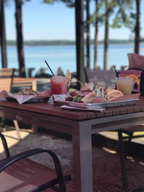 A table with sandwiches, drinks, and snacks, set against a scenic lakeside view with trees in the background.