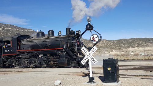 A vintage steam locomotive approaches a railroad crossing with a signal and a black signal box in the foreground.