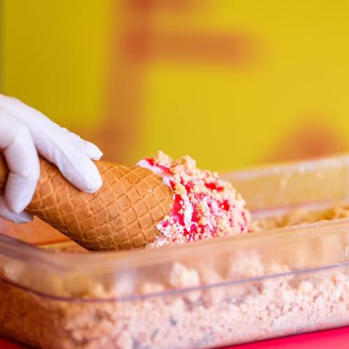 A hand in a white glove dips a waffle cone into a container of ice cream topped with crumbs and sauce.