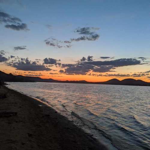 A serene lakeside view at sunset, with colorful clouds reflecting on the water and mountains in the background.