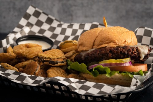 A delicious burger with lettuce, tomato, and onion, served with crispy fried pickles and a dipping sauce on a checkered tray.