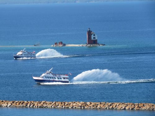 Two boats create waves as they navigate a blue lake near a small red lighthouse on an island.