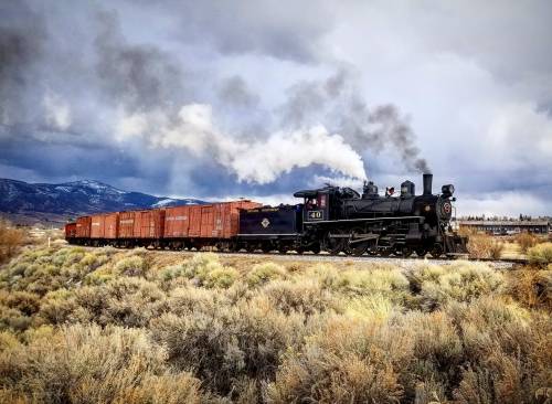 A vintage steam locomotive pulls a line of freight cars through a scenic landscape with mountains and cloudy skies.