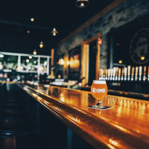 A close-up of a beer glass on a polished bar, with a dimly lit brewery in the background.