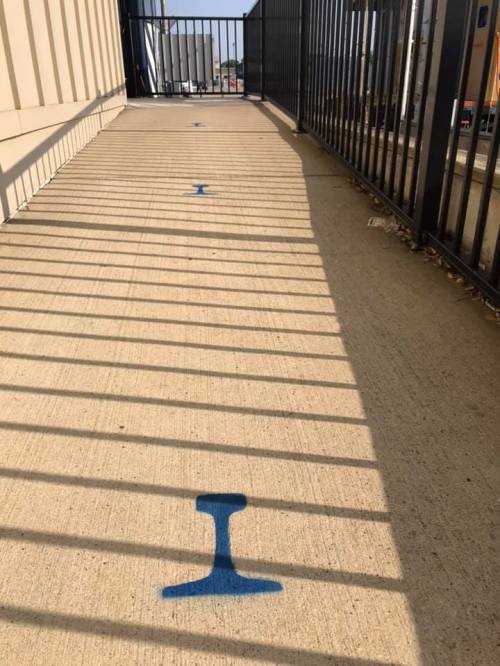 A concrete walkway with blue markings and shadows cast by a fence, leading towards a gate or entrance.
