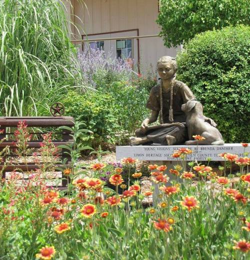 A bronze statue of a girl sitting with a dog, surrounded by colorful flowers and greenery in a garden setting.