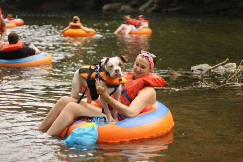 A woman in an orange life jacket sits on an inflatable tube in a river, holding a dog in a life vest.