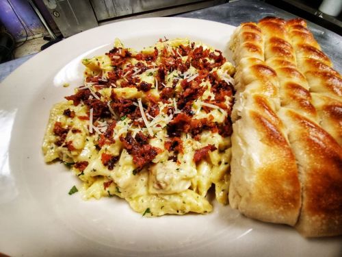 A plate of creamy pasta topped with crispy bacon bits and grated cheese, served with a side of golden breadsticks.