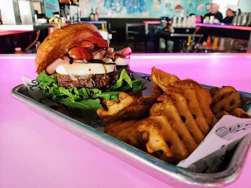 A gourmet burger with toppings on a bed of greens, served with crispy waffle fries on a vibrant pink table.