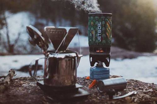 A portable camping stove with steam rising, next to a metal kettle, set in a snowy outdoor environment.