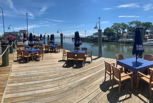 Outdoor dining area with wooden tables and chairs overlooking a calm river on a sunny day. Umbrellas provide shade.