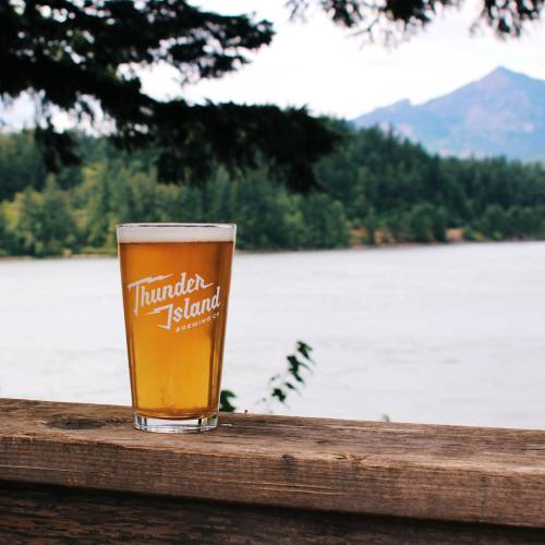 A glass of beer from Thunder Island Brewing sits on a wooden railing with a scenic river and mountains in the background.