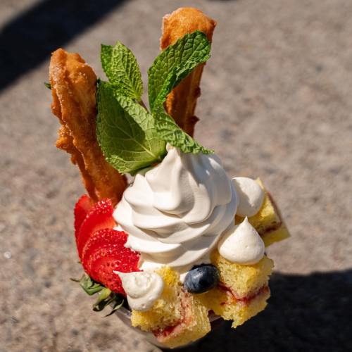 A dessert cup featuring soft-serve ice cream, strawberries, blueberries, cake pieces, and churros, garnished with mint leaves.