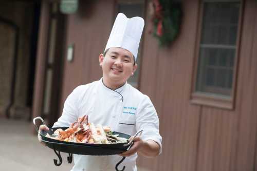 A smiling chef in a white uniform and hat holds a large tray of seafood outside a rustic building.