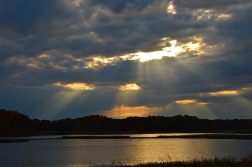 Sunset over a calm lake, with rays of sunlight breaking through dark clouds and reflecting on the water's surface.