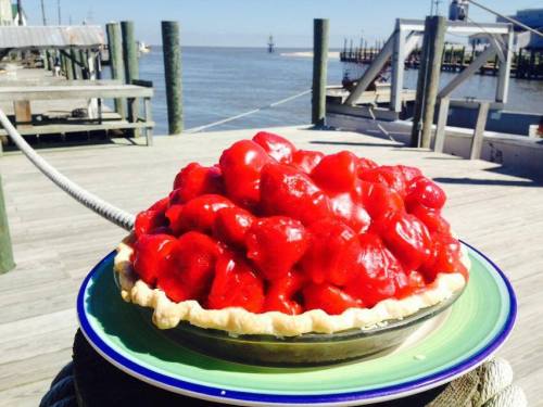 A vibrant strawberry pie sits on a plate by the water, with a wooden dock in the background.