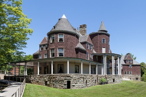 A large, historic mansion with a unique design, featuring a mix of stone and wood, surrounded by green grass and trees.